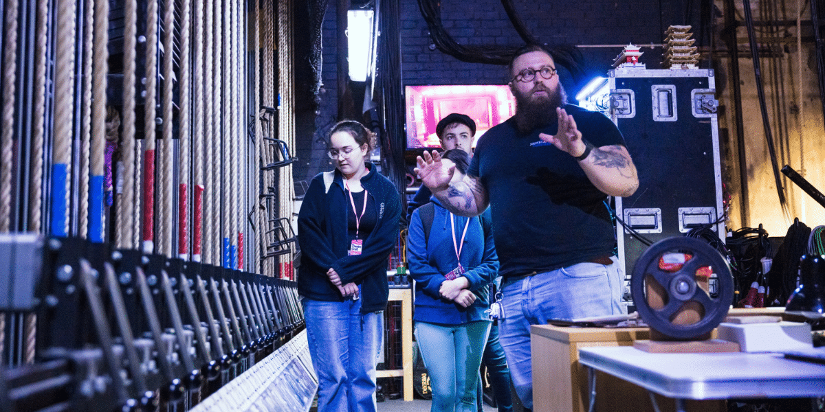 Stage crew members stand backstage among theatre rigging, cables, equipment cases, and lighting gear.