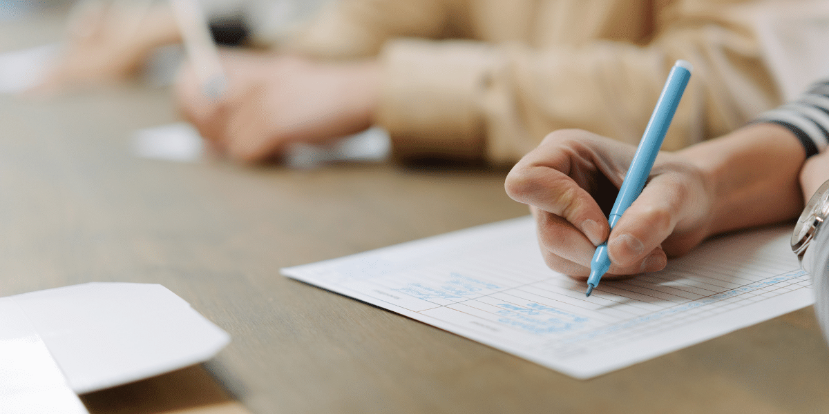 Close-up of a person writing with a blue pen on a lined worksheet at a table, with another person working in the background.
