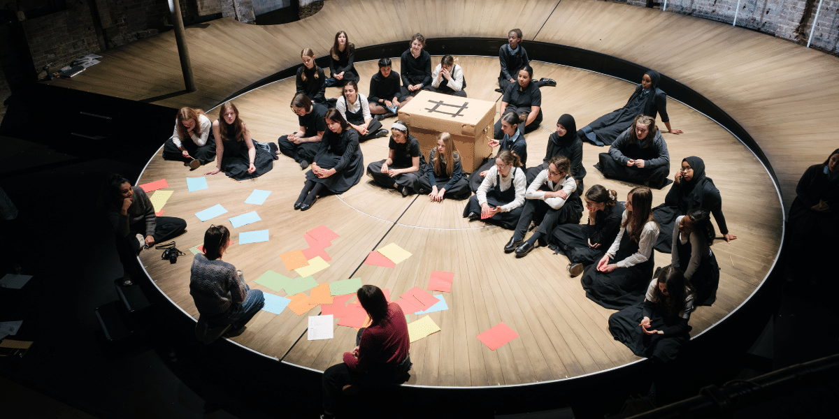 A group of people seated in a circular wooden theatre space, surrounded by colourful paper cards and a large cardboard box.
