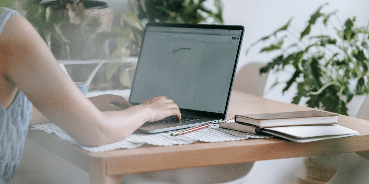 A person sat at a wooden table typing notes onto a laptop. On the desk there are various items of stationery. The blurred background features multiple green plants.