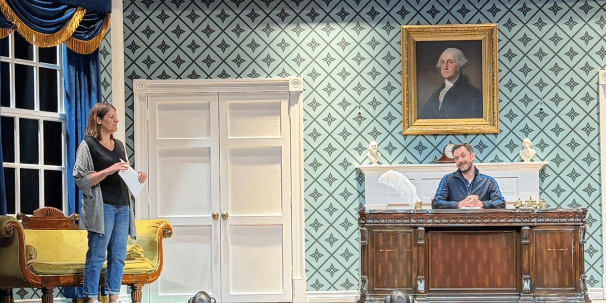 Woman stands beside a couch while a man sits behind an antique desk in a patterned, historic-looking room with a George Washington portrait.