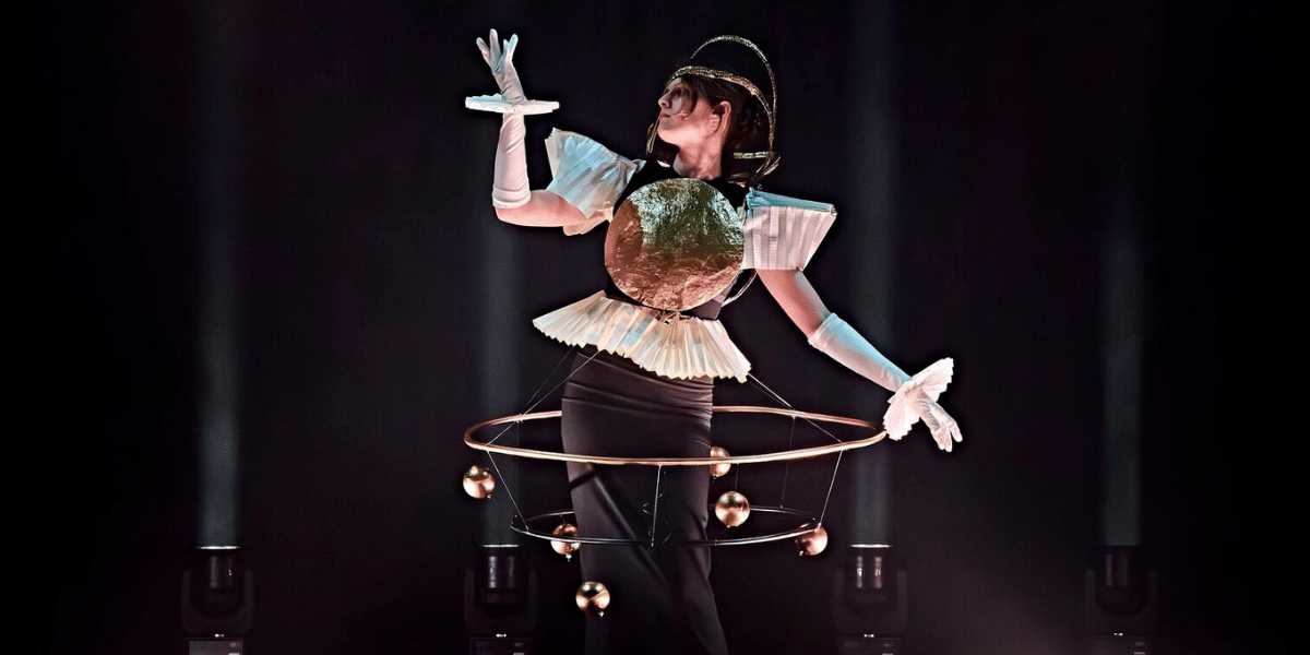 Performer in a futuristic planetary costume with metallic chest plate, orbiting rings, and floating spheres on a dark stage.