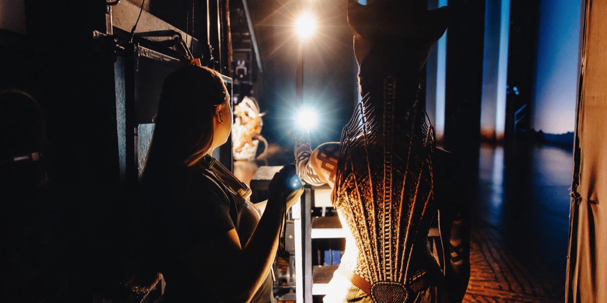 Backstage view of a performer in an elaborate beaded costume and a crew member preparing under bright stage lights.