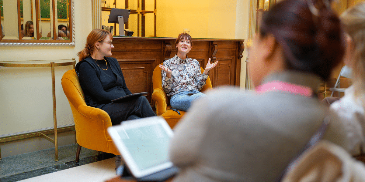 Two people engage in conversation in a cosy setting, with one gesturing expressively while seated in yellow armchairs.