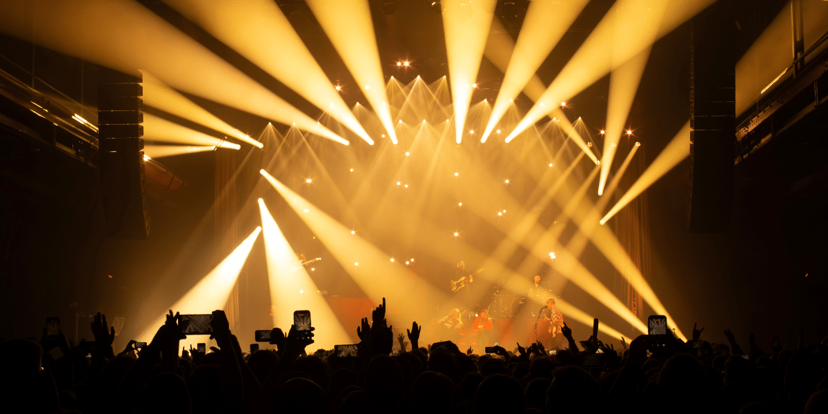 Concert stage illuminated by golden beams, with musicians performing and a silhouetted crowd recording on smartphones.