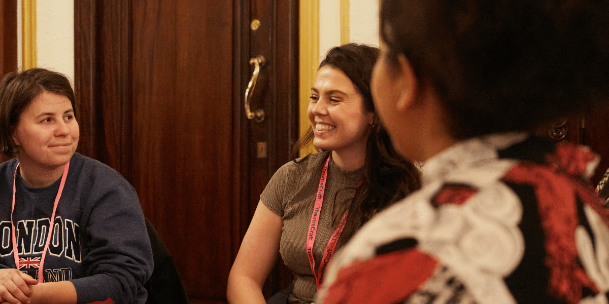 A group of people engage in a discussion at a table, with a wooden door in the background and colourful lanyards visible around their necks.
