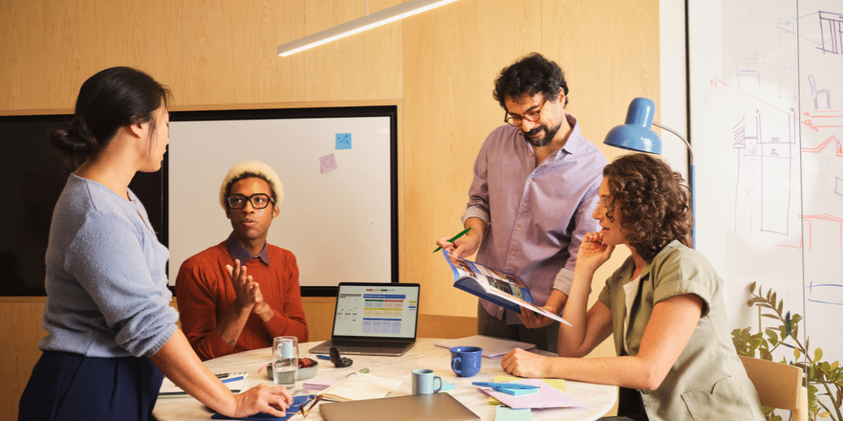 Four coworkers collaborate around a table in a bright office, discussing documents beside a laptop and whiteboard sketches.