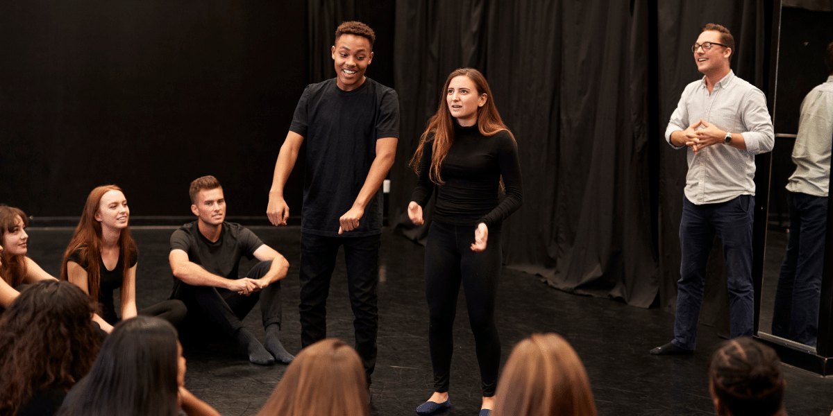 Actors rehearse on a black-box theatre stage while seated performers watch.