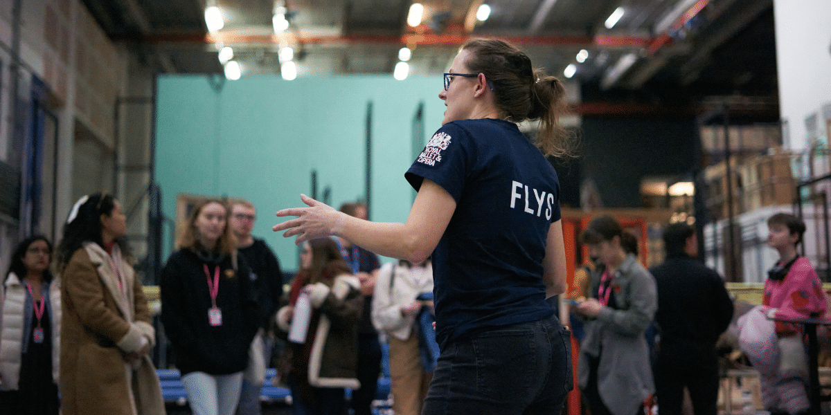 A person in a "FLYS" shirt stands with their back turned, addressing a group of engaged individuals in an industrial setting.