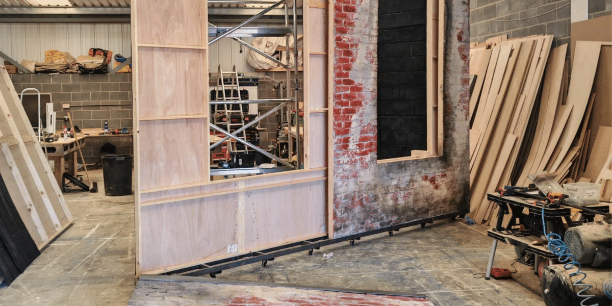 Workshop with a partially built timber and exposed-brick wall, scaffolding, tools, lumber, and construction materials.