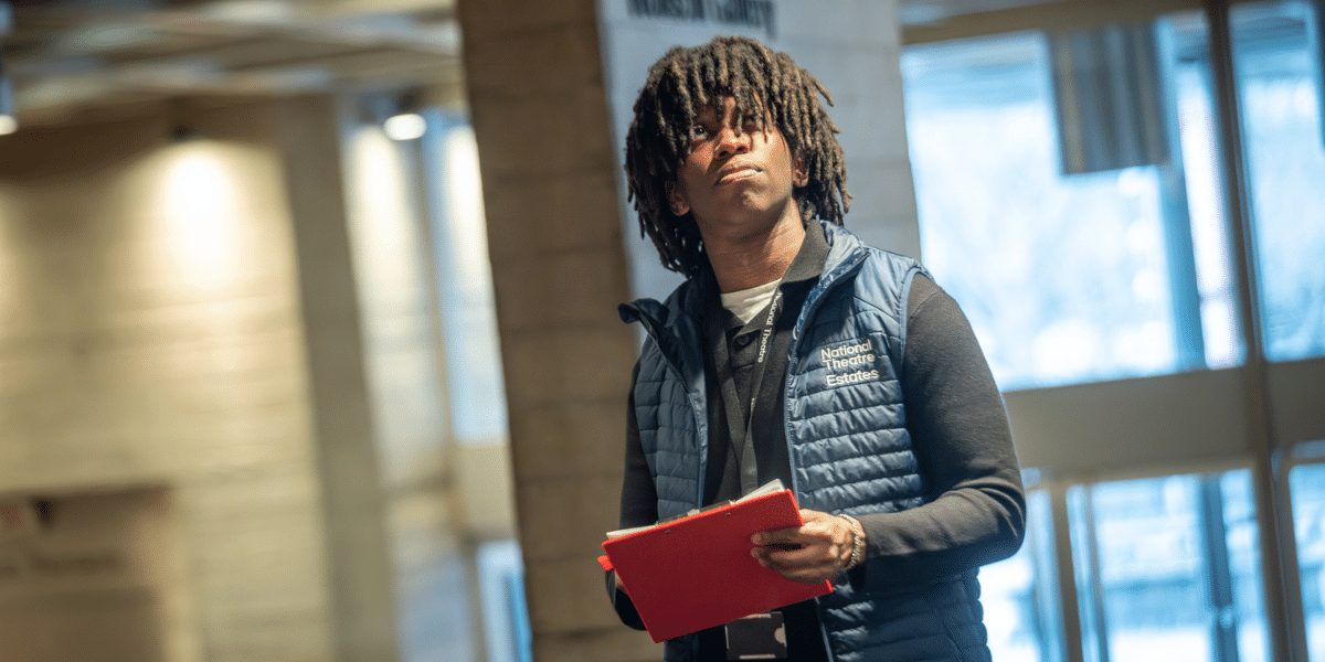 A young man with dreadlocks wearing a National Theatre Estates vest holds a red folder inside a bright building.