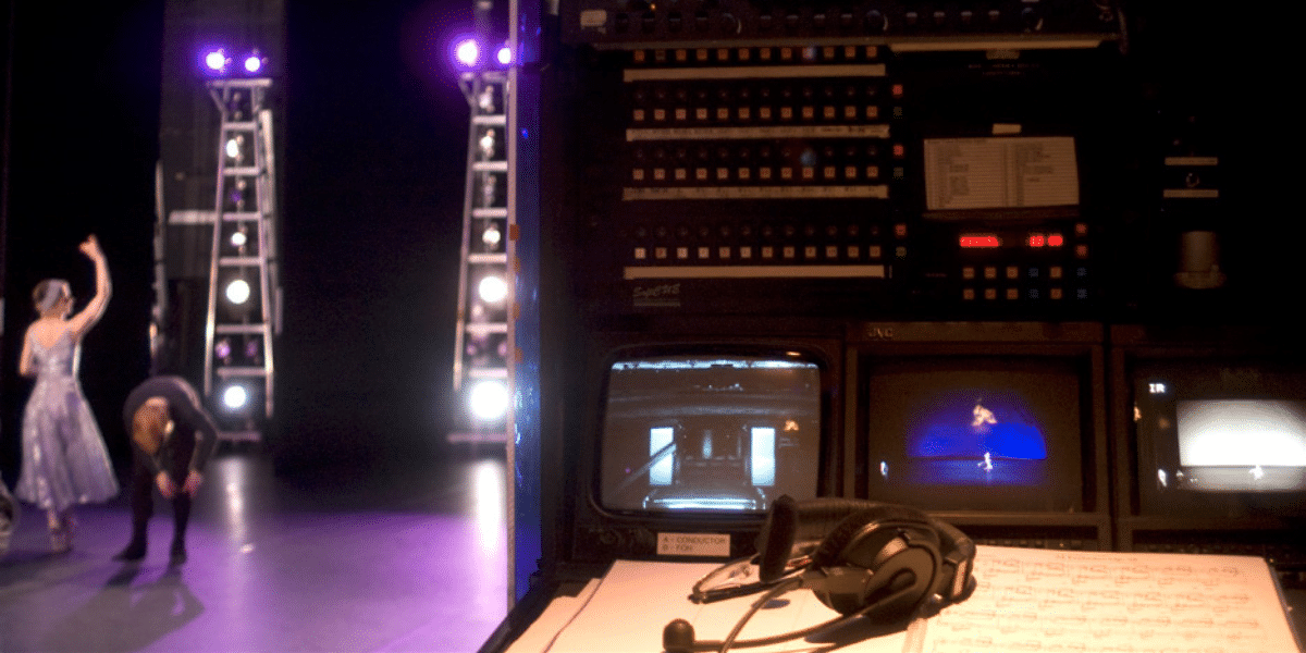 Backstage view of a theatre with lighting equipment, monitors, a script, and a headset, with dancers on stage in the background.