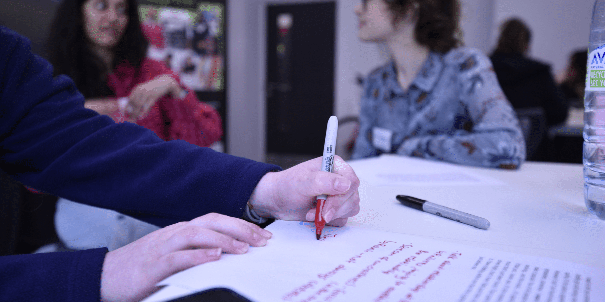 Person writes on a large sheet of paper with a red marker at a meeting table, while others sit nearby.