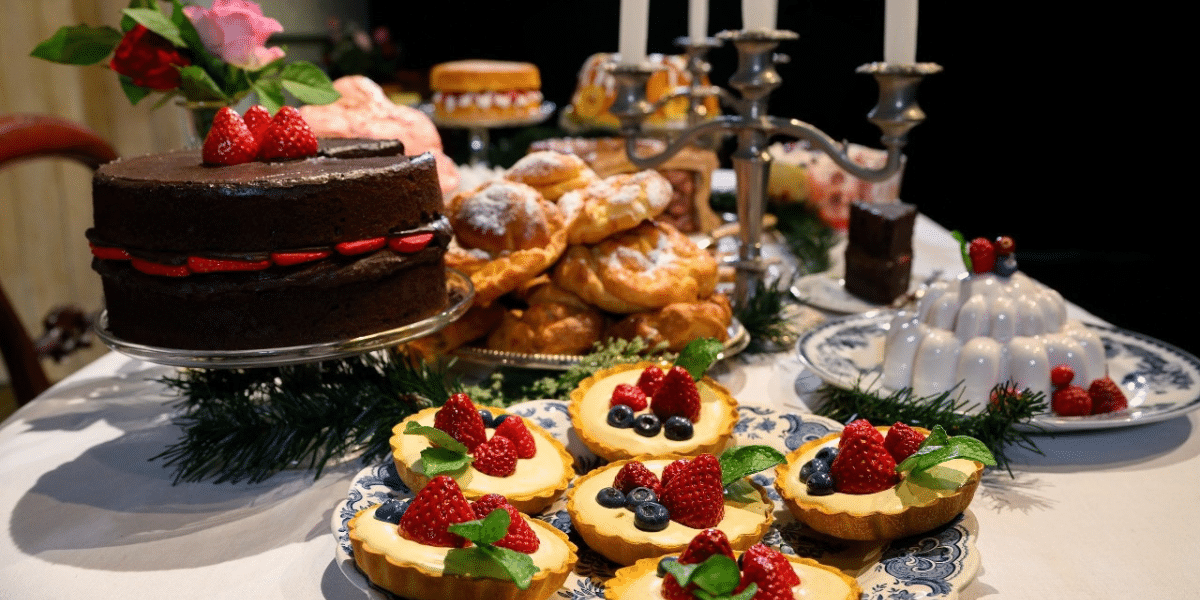 Dessert table featuring props of chocolate cake, berry-topped fruit tarts, pastries, pudding, flowers, and festive greenery.