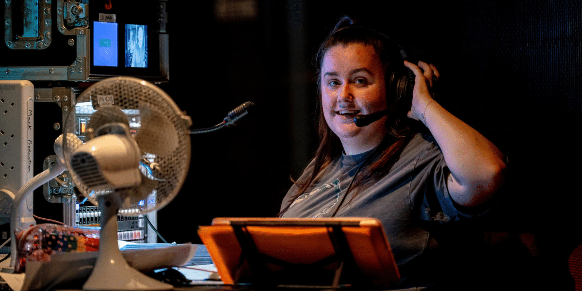 Woman wearing a headset in a dim control room, seated behind monitors and a desk fan.