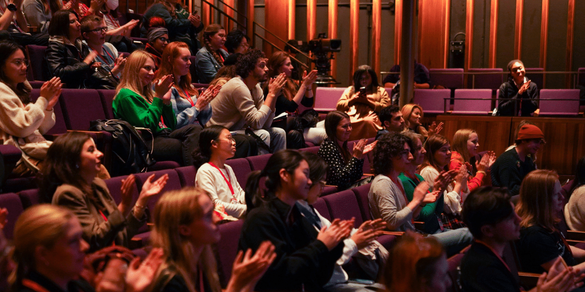Audience applauding in a warmly lit auditorium with tiered purple seats.