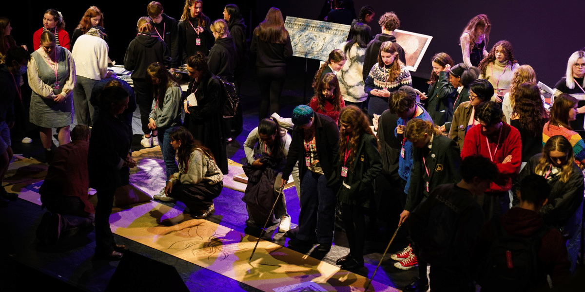 A diverse group of people engages in the Painting Worlds workshop, surrounded by various artistic displays on stage and using long paint sticks to draw.