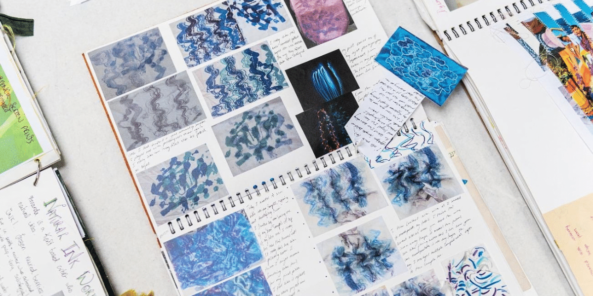 Open sketchbook displaying blue abstract textile pattern studies, handwritten notes, and colorful reference images on a white table.