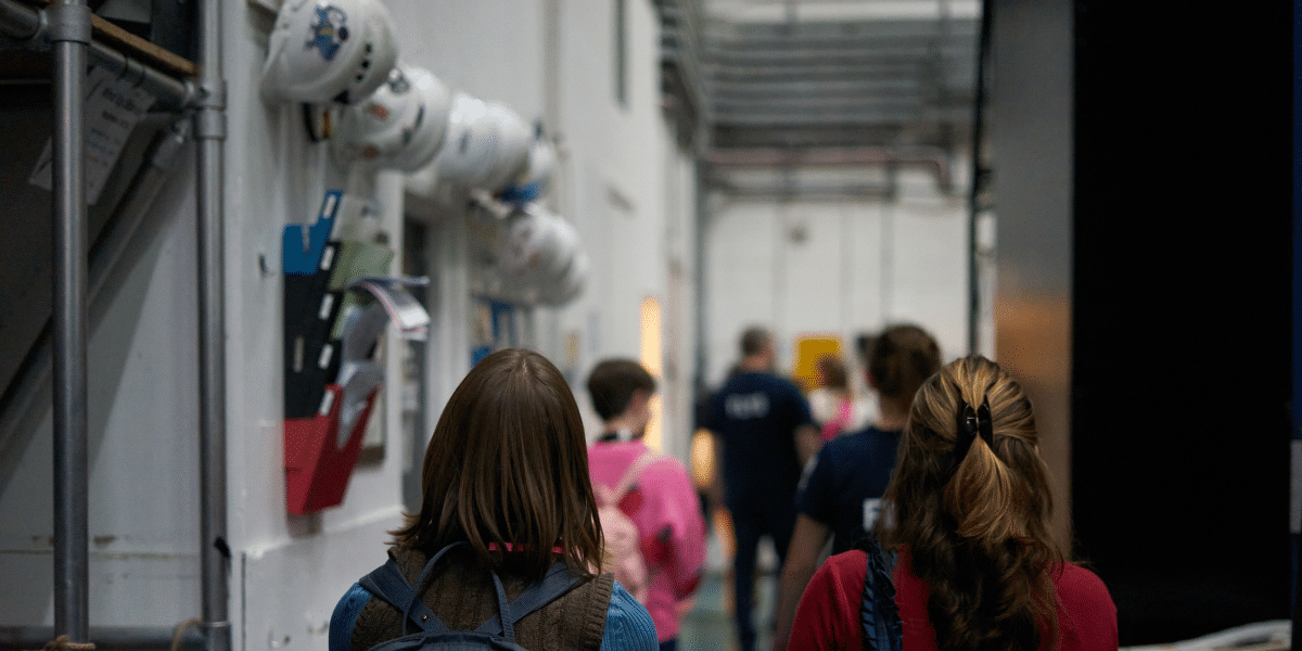 A group of people walk down a hallway lined with hard hats mounted on the wall.