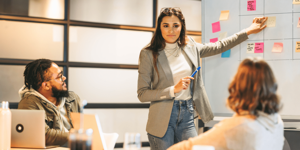 A person presents ideas using a whiteboard and sticky notes during a collaborative meeting, while two colleagues listen attentively.