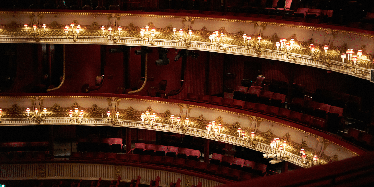 Ornate, empty balconies inside the Royal Opera House theatre, with red seats and glowing lamps.