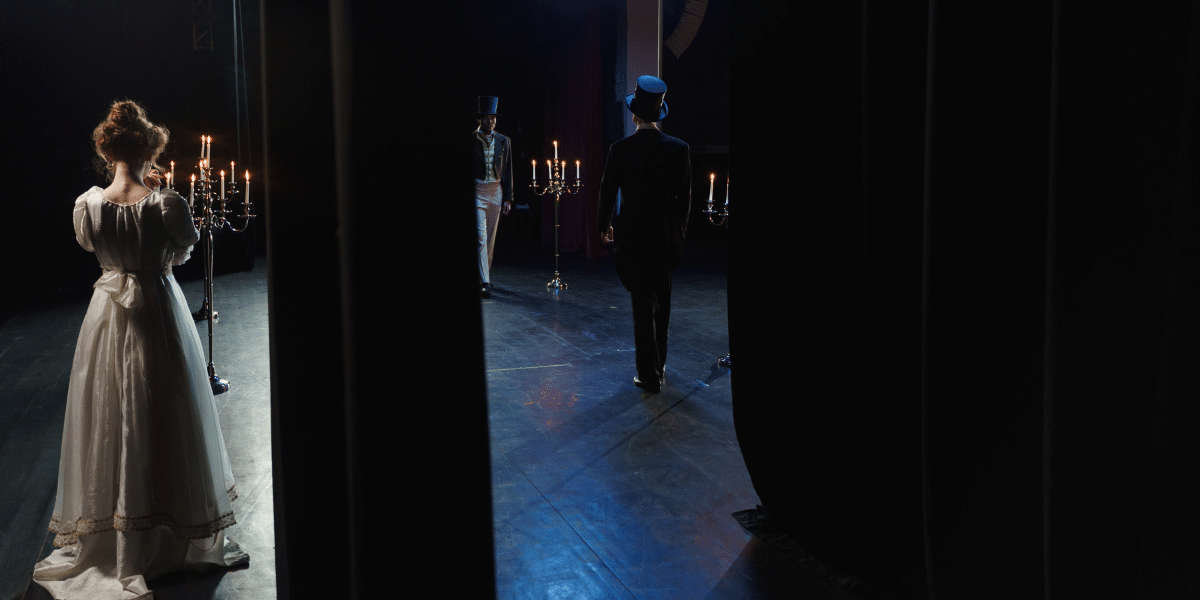Period-costumed actors stand among candlelit candelabras on a dark theater stage, viewed through curtains.