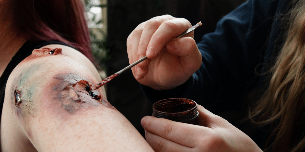 A woman skillfully creates the illusion of an open wound, applying SFX makeup and prosthetics for a realistic effect.