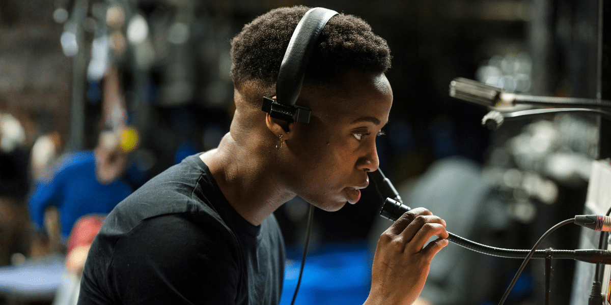 A young person wearing headphones speaks into a microphone in a dimly lit backstage theatre setting.