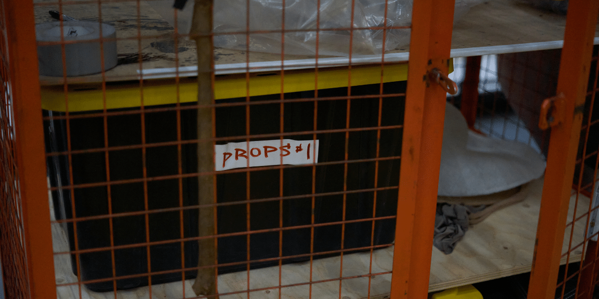 A black storage bin labeled "PROPS #1" inside an orange metal cage, with various items peeking out from behind it.