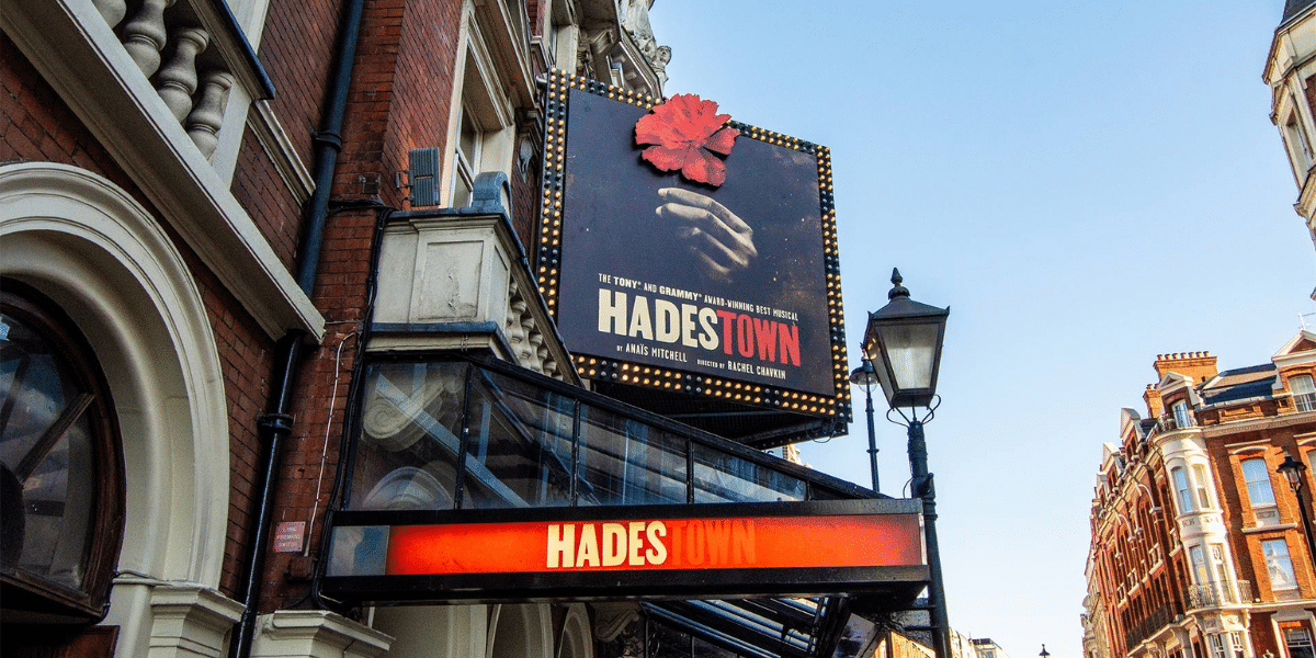 Hadestown musical plaque mounted on a historic red-brick theatre, with ornate buildings and a clear blue sky.