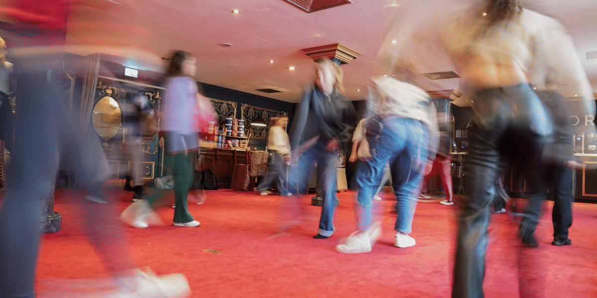 Motion-blurred people walk across a red-carpeted, ornate theatre lobby.