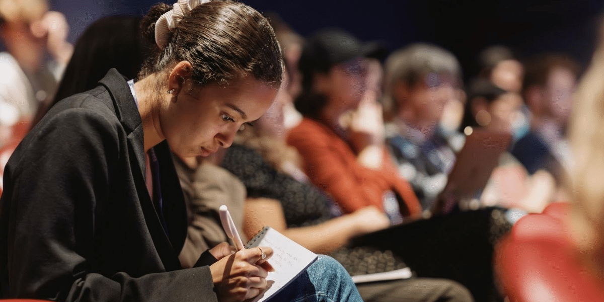 A group of people sat down in an auditorium with attentive facial expressions. The background is blurred and the camera is focused on a person writing notes into a notebook.