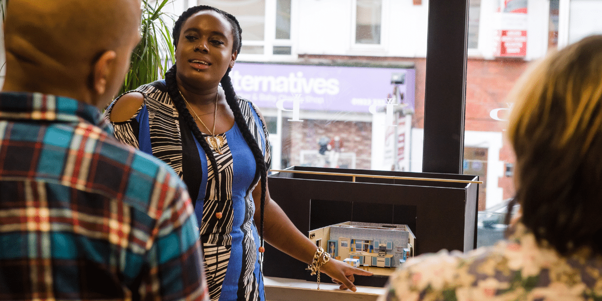 Woman presents a miniature architectural model to two people in a bright indoor setting.
