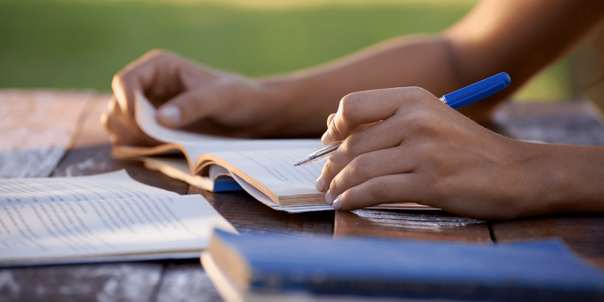 Person writing with a blue pen in an open notebook on a wooden table, with books nearby.